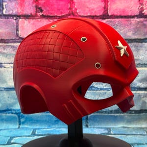 May include: A bright red helmet with a star emblem on the side. The helmet has a woven texture on the top and several small silver rivets. The eye opening is large and the helmet is on a black stand.