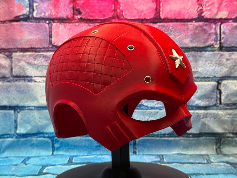 May include: A bright red helmet with a star emblem on the side. The helmet has a woven texture on the top and several small silver rivets. The eye opening is large and the helmet is on a black stand.