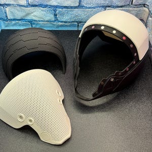 May include: Three-piece protective helmet set. Includes a white helmet with a black trim and a black helmet piece. The third piece is a perforated off-white face shield with circular openings.
