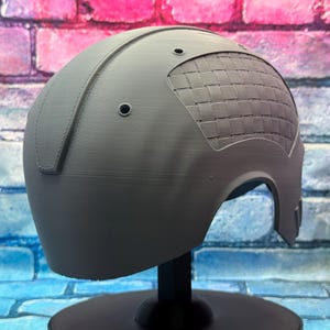 May include: A gray helmet with a woven pattern on the upper right side. The helmet has a smooth, matte finish and is mounted on a black stand. Two small holes are visible near the top.