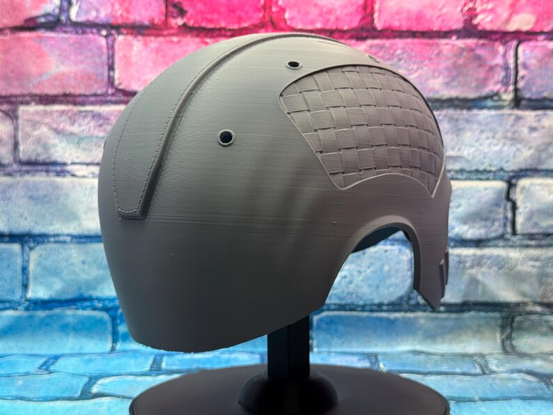May include: A gray helmet with a woven pattern on the upper right side. The helmet has a smooth, matte finish and is mounted on a black stand. Two small holes are visible near the top.