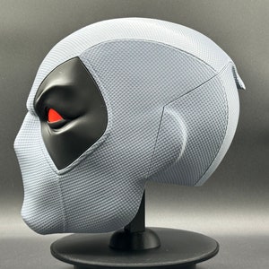 Może przedstawiać: Szara maska Deadpoola z czarnym okiem i czerwonym detalem oka. Maska stoi na czarnym stojaku.