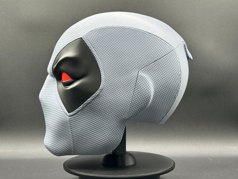 Może przedstawiać: Szara maska Deadpoola z czarnym okiem i czerwonym detalem oka. Maska stoi na czarnym stojaku.