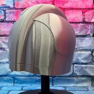 May include: A silver-colored helmet with a textured surface, displayed on a black stand. The helmet has a rounded top and angular side panels. The background is a brick wall with blue, pink, and purple paint.