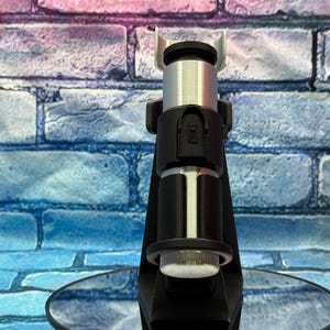 May include: A black and silver lightsaber hilt, with a cylindrical design. The hilt features a black handle with silver accents and a silver emitter. The lightsaber is displayed on a reflective surface against a brick wall background.