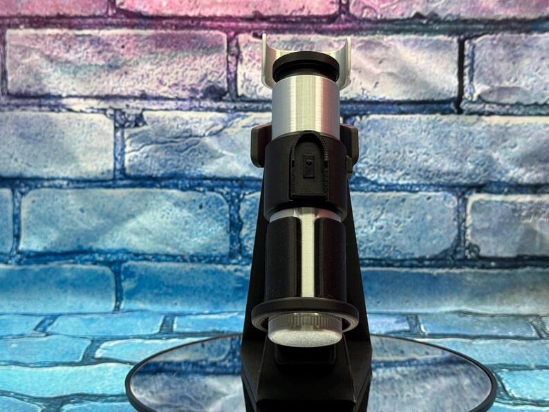 May include: A black and silver lightsaber hilt, with a cylindrical design. The hilt features a black handle with silver accents and a silver emitter. The lightsaber is displayed on a reflective surface against a brick wall background.