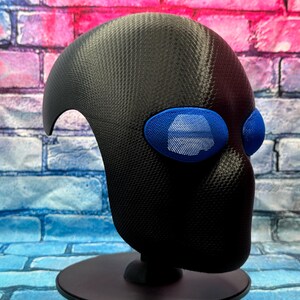 May include: A black full-head mask featuring a textured carbon fiber-like finish and prominent, oval-shaped blue eye lenses with a fine mesh grid pattern. The mask is mounted on a black circular stand, showcasing a smooth, rounded silhouette.