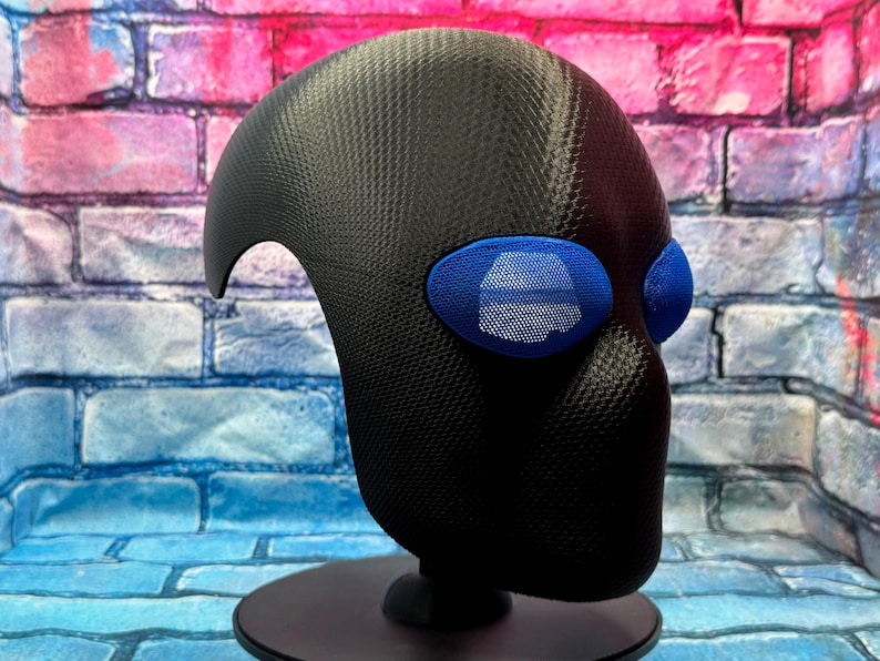 May include: A black full-head mask featuring a textured carbon fiber-like finish and prominent, oval-shaped blue eye lenses with a fine mesh grid pattern. The mask is mounted on a black circular stand, showcasing a smooth, rounded silhouette.