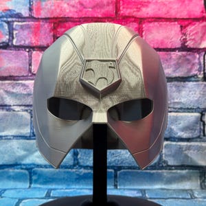 May include: A silver and red superhero mask with a stylized emblem on the forehead. The mask has eye openings and is mounted on a black stand. The background is a brick wall with blue, pink, and purple paint.