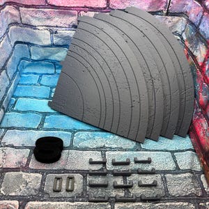 May include: A collection of gray, curved, segmented pieces, possibly for a diorama or model, with a textured surface. Also included are black straps and several small gray rectangular and curved pieces. The items are arranged on a brick-patterned surface.