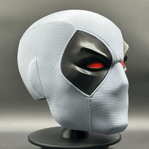 Może przedstawiać: Biała maska Deadpoola z czarnymi otworami na oczy i czerwonymi oczami. Maska wykonana jest z fakturowanego materiału i stoi na czarnym stojaku.