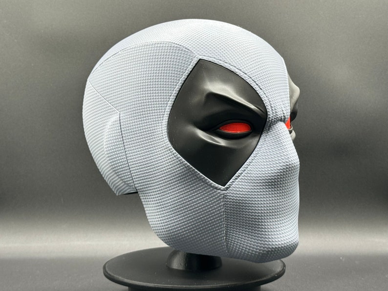 Może przedstawiać: Biała maska Deadpoola z czarnymi otworami na oczy i czerwonymi oczami. Maska wykonana jest z fakturowanego materiału i stoi na czarnym stojaku.