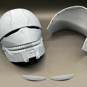 May include: A white plastic helmet with a textured surface and a detailed design. The helmet is in two pieces, with a separate top section. There are two small white plastic pieces that appear to be part of the helmet.