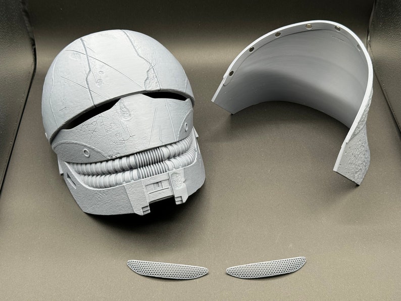 May include: A white plastic helmet with a textured surface and a detailed design. The helmet is in two pieces, with a separate top section. There are two small white plastic pieces that appear to be part of the helmet.