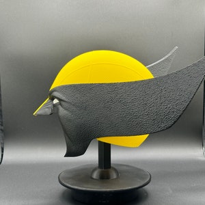 Puede incluir: Una m&aacute;scara 3D impresa en amarillo y negro de un personaje de superh&eacute;roe en un soporte negro. La m&aacute;scara tiene una superficie texturizada y est&aacute; dise&ntilde;ada para parecerse a la cara de un personaje.
