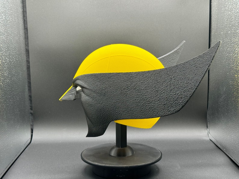 Puede incluir: Una m&aacute;scara 3D impresa en amarillo y negro de un personaje de superh&eacute;roe en un soporte negro. La m&aacute;scara tiene una superficie texturizada y est&aacute; dise&ntilde;ada para parecerse a la cara de un personaje.