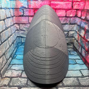 May include: A gray, teardrop-shaped sculpture with a textured surface. The sculpture is divided vertically into two halves, each with a slightly different shade of gray. The design includes curved lines and circular patterns.
