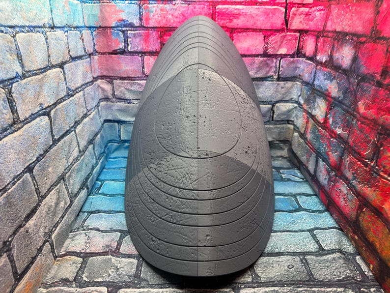 May include: A gray, teardrop-shaped sculpture with a textured surface. The sculpture is divided vertically into two halves, each with a slightly different shade of gray. The design includes curved lines and circular patterns.