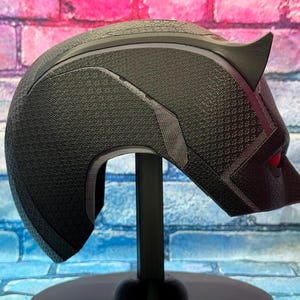 Puede incluir: Un casco negro con una superficie texturizada, con una oreja puntiaguda y detalles de ojos rojos. El casco se muestra sobre un soporte negro sobre un fondo de pared de ladrillos de colores. El dise&ntilde;o recuerda a una m&aacute;scara de superh&eacute;roe.