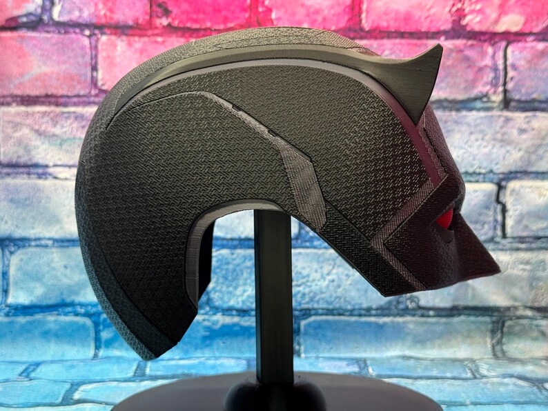 Puede incluir: Un casco negro con una superficie texturizada, con una oreja puntiaguda y detalles de ojos rojos. El casco se muestra sobre un soporte negro sobre un fondo de pared de ladrillos de colores. El dise&ntilde;o recuerda a una m&aacute;scara de superh&eacute;roe.