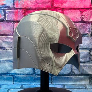 May include: A silver and red helmet with a textured surface, displayed on a black stand. The helmet features a stylized design with a prominent emblem on the side. The background is a brick wall.