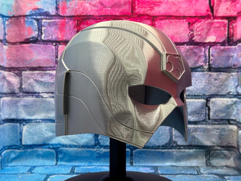 May include: A silver and red helmet with a textured surface, displayed on a black stand. The helmet features a stylized design with a prominent emblem on the side. The background is a brick wall.