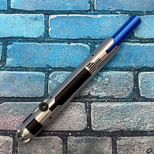 Könnte beinhalten: Ein silberner und schwarzer Stift mit blauer Spitze, der ein schlankes Design mit einer Reihe schwarzer Linien und einem kreisförmigen Knopf aufweist. Der Stift befindet sich vor einem blauen Ziegelhintergrund.