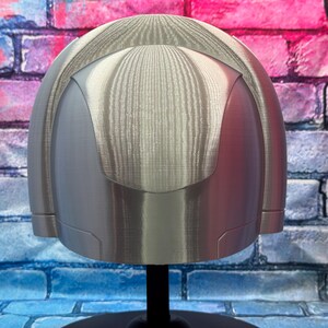 May include: A silver-colored helmet with a rounded top and a smooth, metallic finish. The helmet has a visor-like design and is mounted on a black stand. The background is a brick wall with blue, pink, and purple paint.
