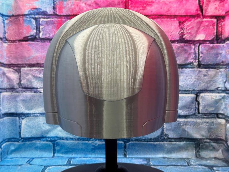 May include: A silver-colored helmet with a rounded top and a smooth, metallic finish. The helmet has a visor-like design and is mounted on a black stand. The background is a brick wall with blue, pink, and purple paint.
