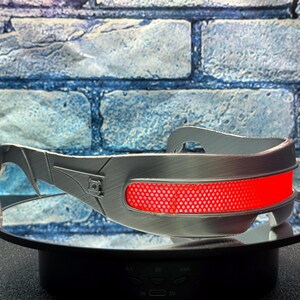 May include: Silver-colored, futuristic-style eyewear with a prominent red lens. The glasses have a sleek, metallic design with angular details and a curved top bar. The red lens is a mesh-like material.