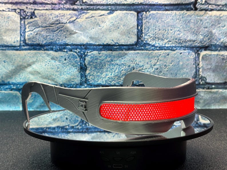 May include: Silver-colored, futuristic-style eyewear with a prominent red lens. The glasses have a sleek, metallic design with angular details and a curved top bar. The red lens is a mesh-like material.