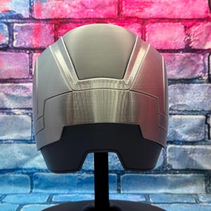 May include: A silver and gray helmet with a brushed metal finish. The helmet has a geometric design with angular panels and a dark gray base. It is displayed on a black stand against a colorful brick wall.