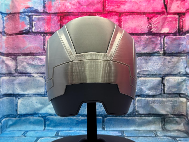 May include: A silver and gray helmet with a brushed metal finish. The helmet has a geometric design with angular panels and a dark gray base. It is displayed on a black stand against a colorful brick wall.