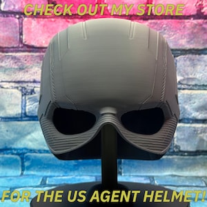May include: A gray US Agent helmet, displayed on a black stand against a brick wall background. The helmet has a detailed design with eye openings and a textured lower section. Text at the top reads "CHECK OUT MY STORE" and at the bottom "FOR THE US AGENT HELMET!"