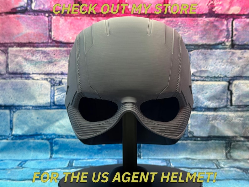 May include: A gray US Agent helmet, displayed on a black stand against a brick wall background. The helmet has a detailed design with eye openings and a textured lower section. Text at the top reads "CHECK OUT MY STORE" and at the bottom "FOR THE US AGENT HELMET!"