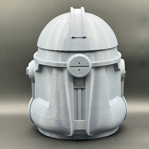 Puede incluir: Un casco gris impreso en 3D con un dise&ntilde;o detallado. El casco tiene una parte superior redondeada y una visera con una peque&ntilde;a abertura. El casco est&aacute; hecho de pl&aacute;stico y tiene una superficie lisa.