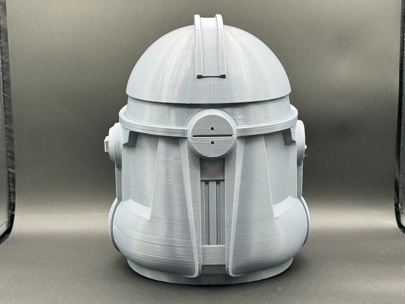 Puede incluir: Un casco gris impreso en 3D con un dise&ntilde;o detallado. El casco tiene una parte superior redondeada y una visera con una peque&ntilde;a abertura. El casco est&aacute; hecho de pl&aacute;stico y tiene una superficie lisa.