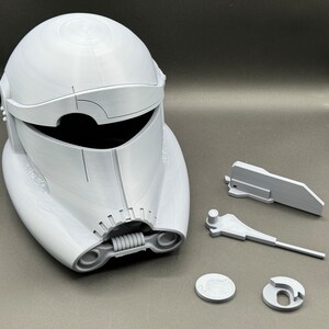 May include: A white plastic helmet with a visor and a spring-loaded jaw. The helmet is disassembled into several pieces, including the helmet itself, the visor, the jaw, and two small circular pieces.