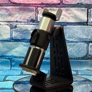 May include: A black and silver lightsaber replica, displayed on a black stand. The lightsaber has a silver hilt and a black handle with intricate details. The stand is triangular with cut-out designs. The background is a brick wall with blue, pink, and purple hues.