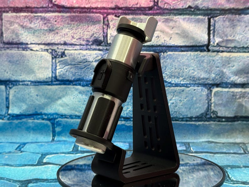May include: A black and silver lightsaber replica, displayed on a black stand. The lightsaber has a silver hilt and a black handle with intricate details. The stand is triangular with cut-out designs. The background is a brick wall with blue, pink, and purple hues.