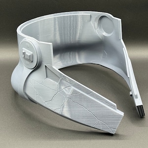 May include: A gray plastic helmet with a textured surface and a small, round button on the side. The helmet is in two pieces, with the front piece separated from the back piece.