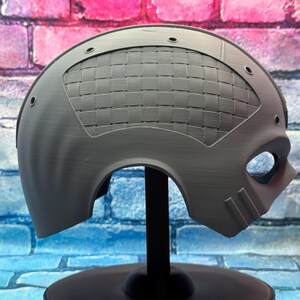 May include: A gray, futuristic-style mask with a woven pattern on the upper portion. The mask has eye and mouth openings, and is mounted on a black stand. The background is a brick wall with blue, pink, and purple colors.