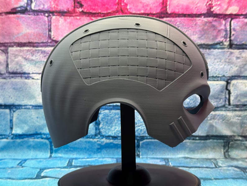 May include: A gray, futuristic-style mask with a woven pattern on the upper portion. The mask has eye and mouth openings, and is mounted on a black stand. The background is a brick wall with blue, pink, and purple colors.