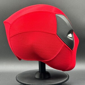 May include: Red and black Deadpool mask with a textured surface. The mask is on a black stand.