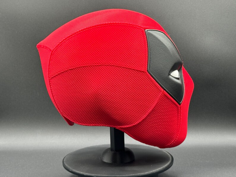 May include: Red and black Deadpool mask with a textured surface. The mask is on a black stand.