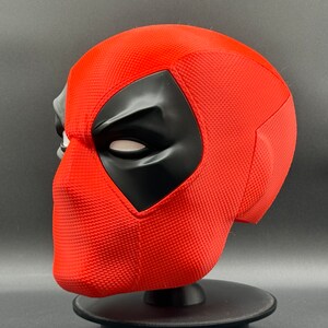 Puede incluir: Una m&aacute;scara de Deadpool roja y negra con una superficie texturizada. La m&aacute;scara est&aacute; en un soporte negro.