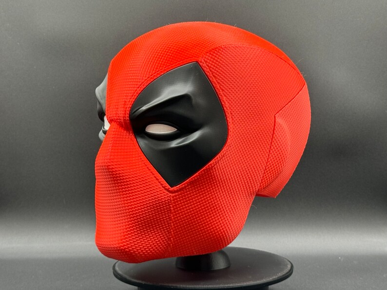 Puede incluir: Una m&aacute;scara de Deadpool roja y negra con una superficie texturizada. La m&aacute;scara est&aacute; en un soporte negro.