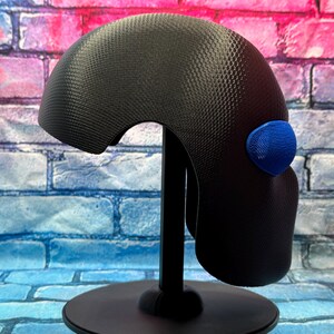 May include: A black helmet with a textured, honeycomb-like surface pattern and a single, prominent blue oval-shaped accent piece on the side. The helmet is mounted on a black display stand, showcasing its curved, rounded profile and smooth edges.