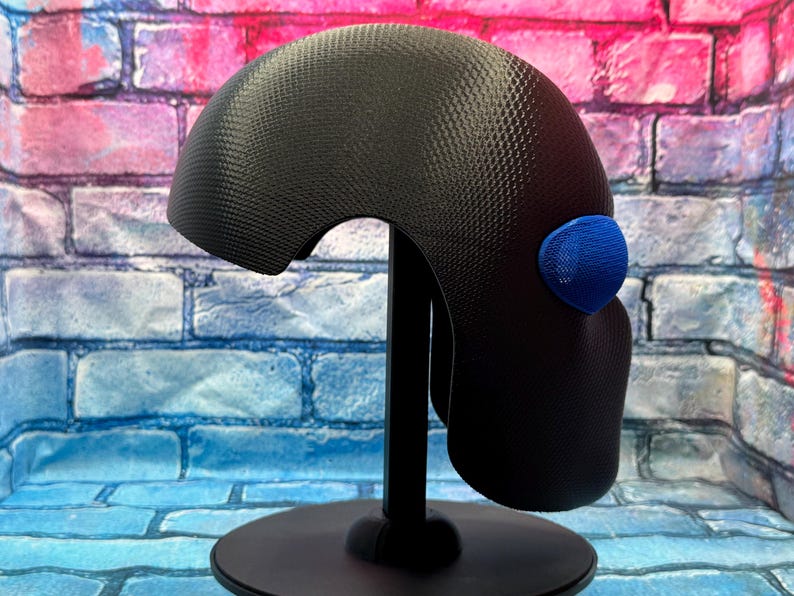 May include: A black helmet with a textured, honeycomb-like surface pattern and a single, prominent blue oval-shaped accent piece on the side. The helmet is mounted on a black display stand, showcasing its curved, rounded profile and smooth edges.