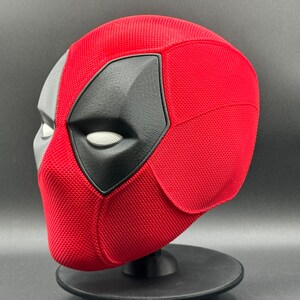 May include: A red and black Deadpool mask with a textured surface. The mask is on a black stand.